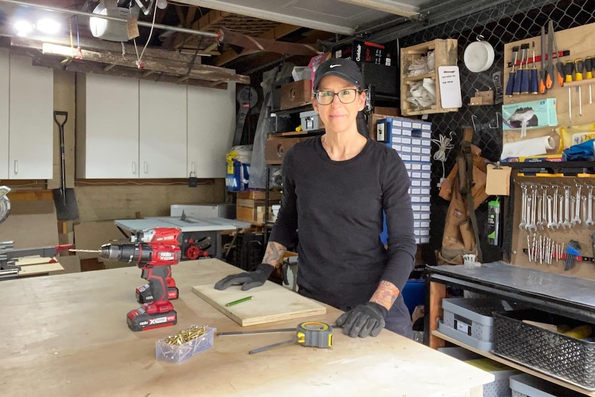 A woman wearing a cap and safety glasses stands at a wooden workbench.