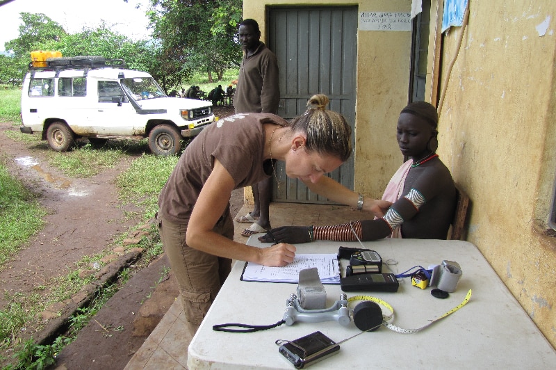 A Nilo-Saharan person is sitting down while an American scientist collects a skin reflectance ready from their arm