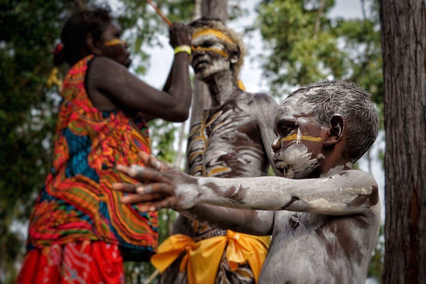 An Aboriginal woman paints Eddie William Gumbula in white and yellow body paint as a young child plays in the foreground.