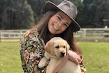 A young woman in a hat holding a golden puppy stands smiling to camera