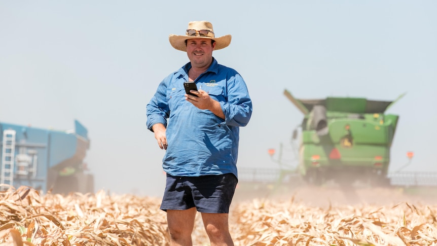 A smiling farmer stands in a paddock, holding his phone.