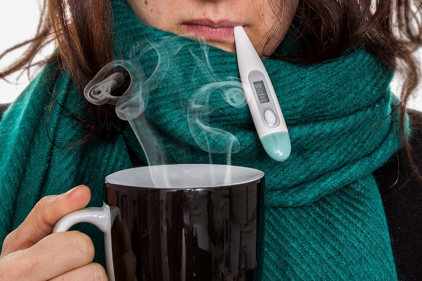 Close-up of a woman with a green scarf around her neck holding a cup of tea and thermometer in her mouth