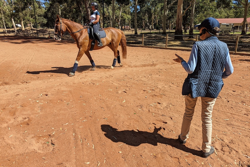 Woman in puffy vest and hat stands in arena talking to woman riding horse