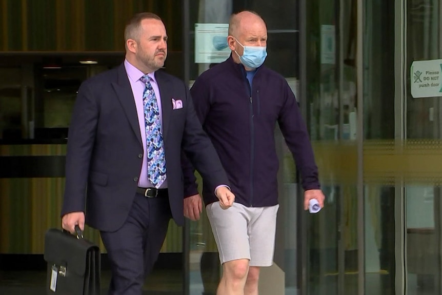 Two men exit the doors of a court building. The lawyer is wearing a suit, the other man is wearing shorts and a jumper.