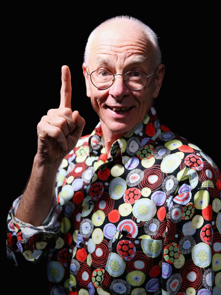 A man wearing a brightly patterened shirt smiles and holds his index finger aloft.