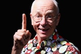 A man wearing a brightly patterened shirt smiles and holds his index finger aloft.