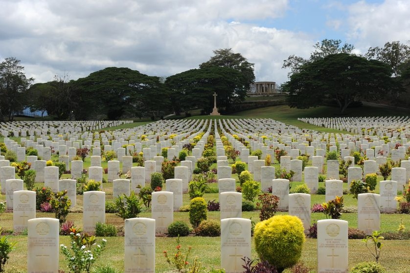 A wide shot shows cemetery dotted with almost identical white gravestones, with a statue of a cross on a mound in the distance