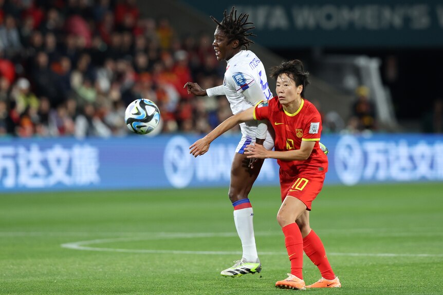 Two female football players, one in white and the other in red, compete for the football which is bouncing away