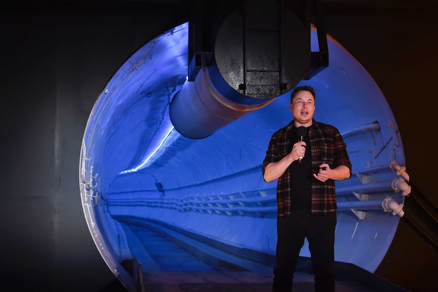 Elon Musk wears a plaid shirt, black t-shirt and black trousers as he speaks at the mouth of a blue-lit tunnel with microphone.