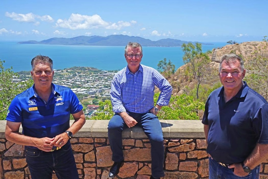 Three men wearing blue shirts smiling at the camera. In the background is an aerial view of Magnetic Island and Townsville.