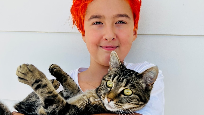 A young boy with orange hair looking at the camera while holding a cat