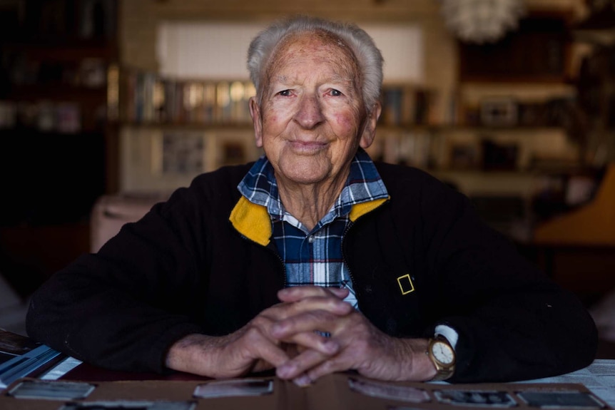 Image of an elderly man sitting at a table, wearing a black and yellow jacket and a checked shirt. His hands are interlocked.
