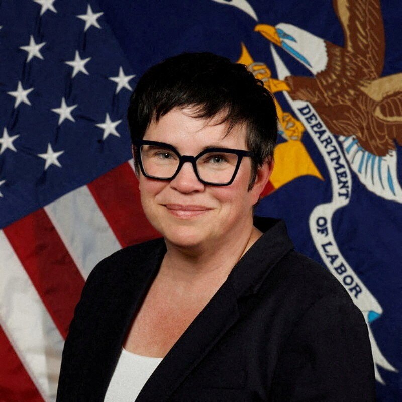 Erika McEntarfer posing for a photograph and grinning in front of a US Department of Labor flag