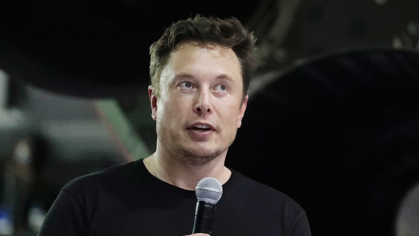 Elon Musk is wearing a black tee-shirt and speaking into a microwave at a public event