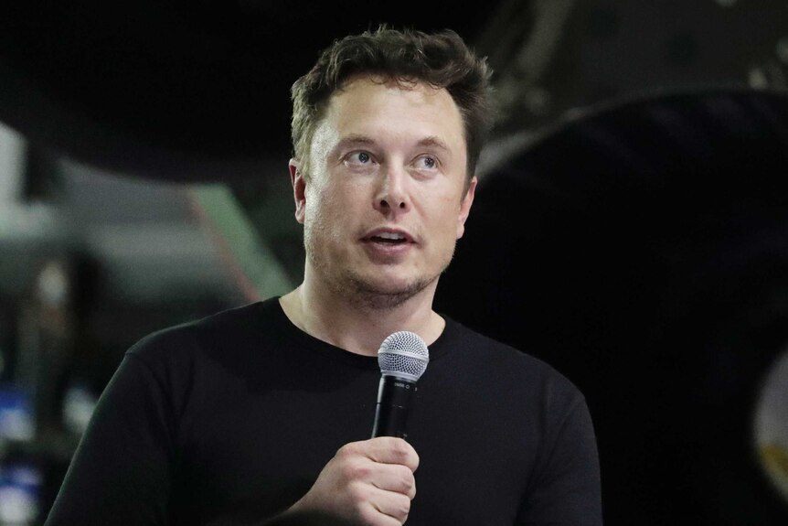 Elon Musk is wearing a black tee-shirt and speaking into a microphone at a public event