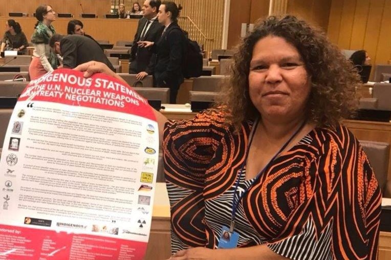 A woman with dark curly hair holds up a document while sitting in a room with people behind her