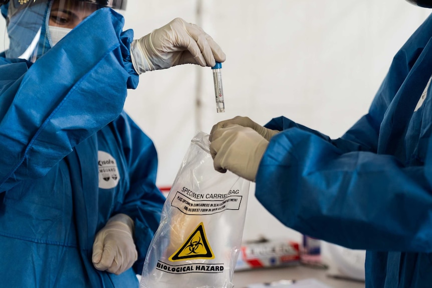 Two testing staff in protective gear lower a COVID-19 swab sample into a bag labelled biological hazard.