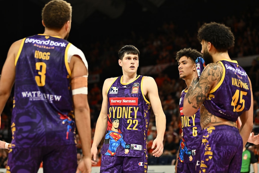 A medium shot of four male basketballers standing near each other during a game, with a crowd behind them