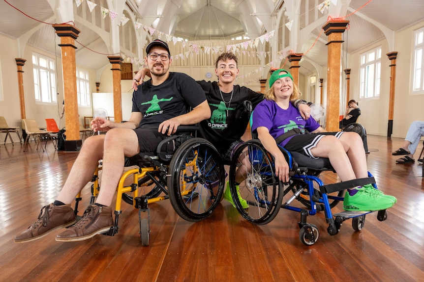 Three people smile and pose for a photo in a dance studio, two people use a wheelchair.