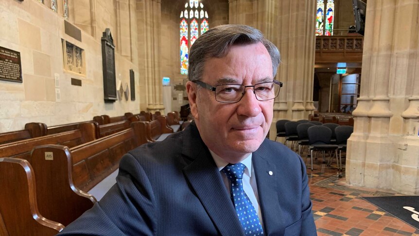 Medium close up of Rev Glenn Davies, Anglican Archbishop of Sydney, sitting on a pew in St Andrew's Cathedral, looking at camera