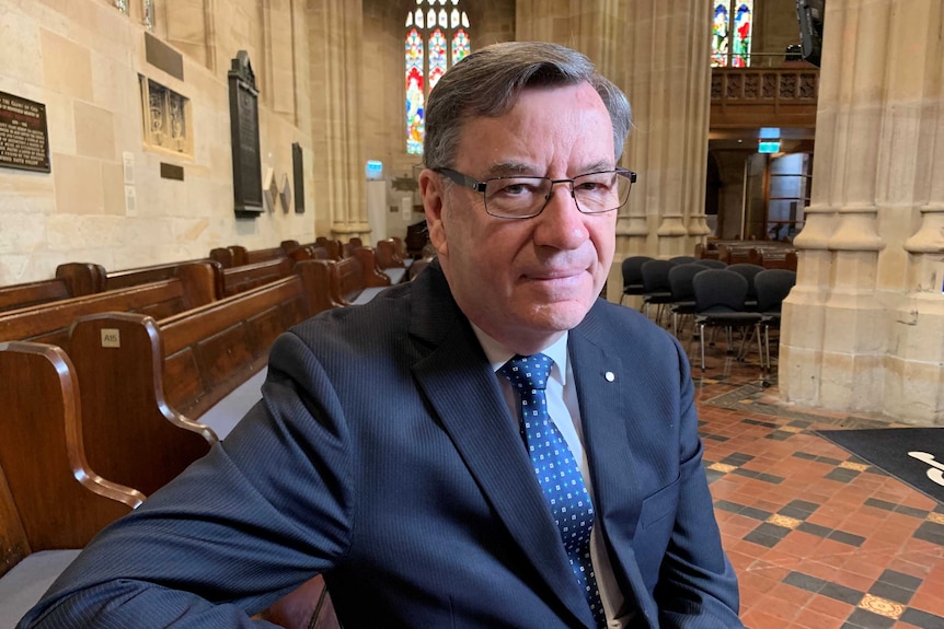 Medium close up of Rev Glenn Davies, Anglican Archbishop of Sydney, sitting on a pew in St Andrew's Cathedral, looking at camera