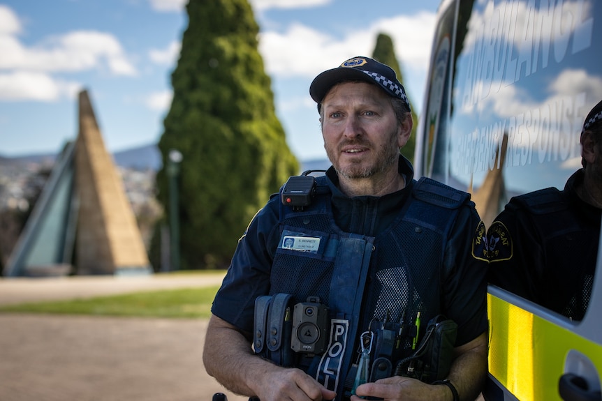 A police officer leans against a police vehicle and smiles at the camera.