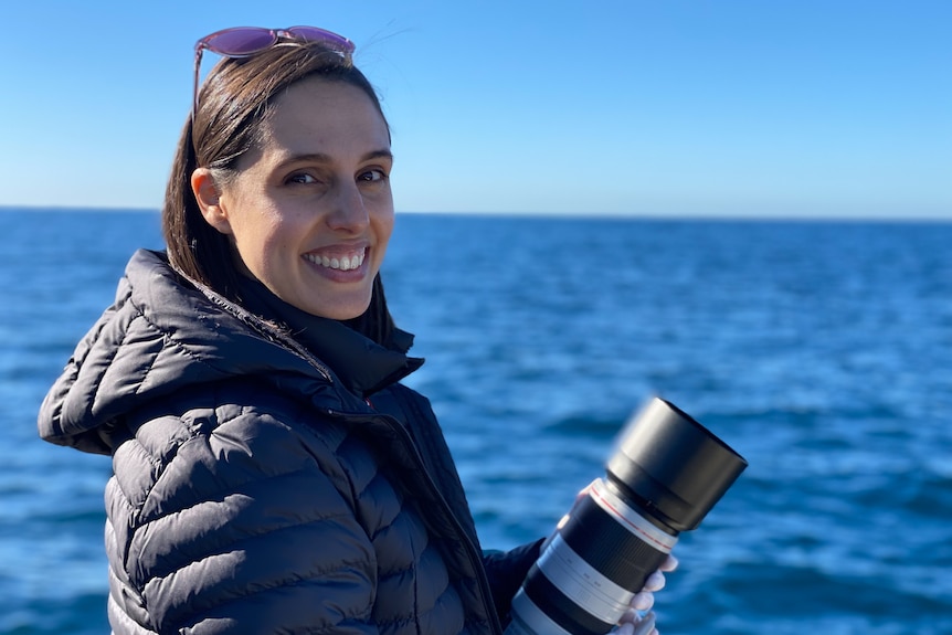 Dr Vanessa Pirotta. woman stands with camera in front of ocean