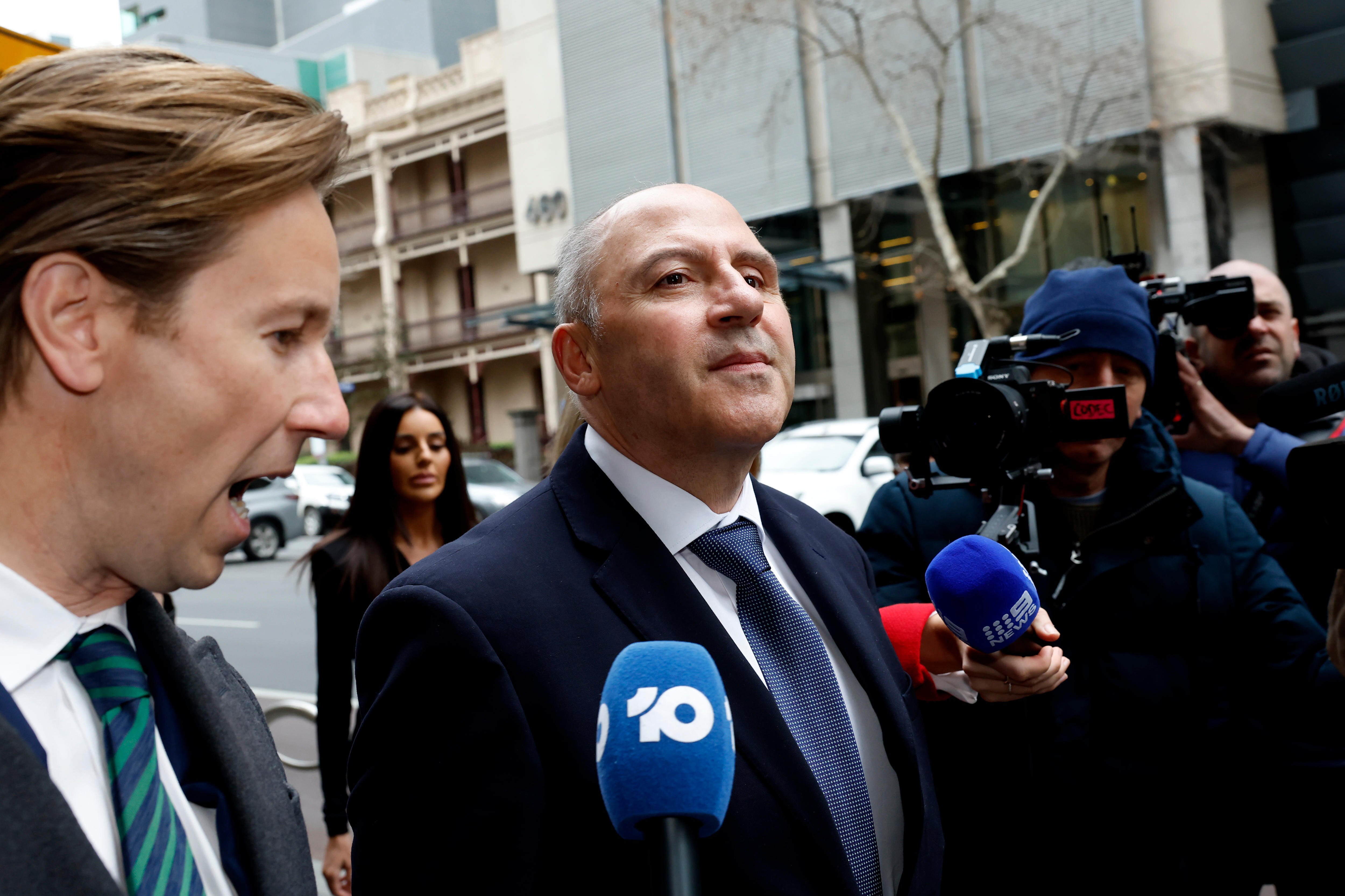 A bald man in a blue suit, white shirt and blue tie walks along a street with tv cameras surrounding him.