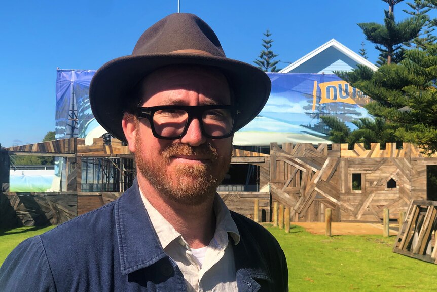 Man wearing glasses and hat smiles in front of wooden structure and festival sign. 