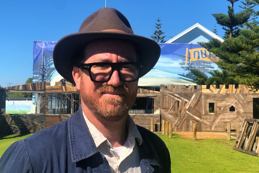 Un homme portant des lunettes et un chapeau sourit devant une structure en bois et un panneau de festival. 