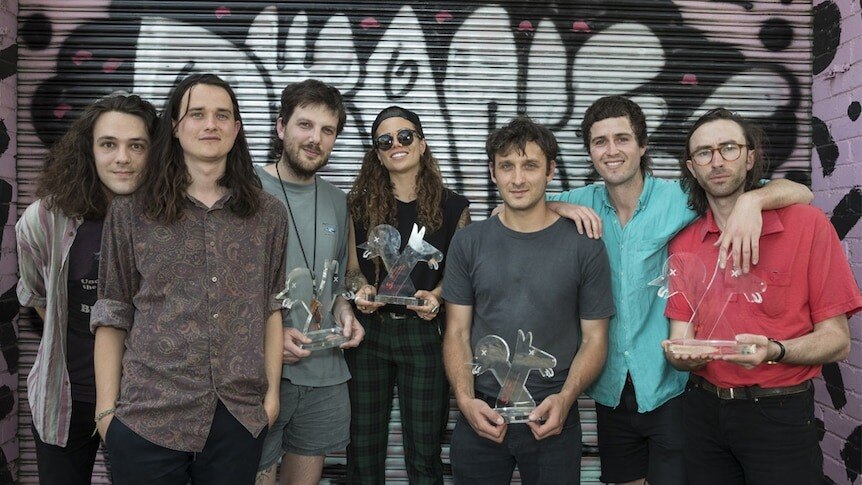 seven people pose against a graffiti&#x27;d garage roller door with transparent J Awards trophies. 