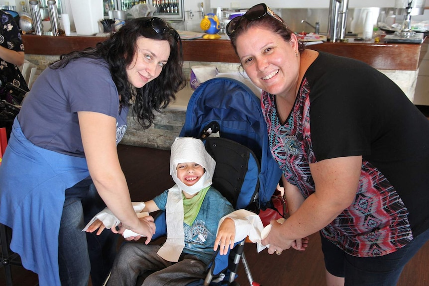 A young boy in a wheelchair is wrapped up in toilet paper by two women.
