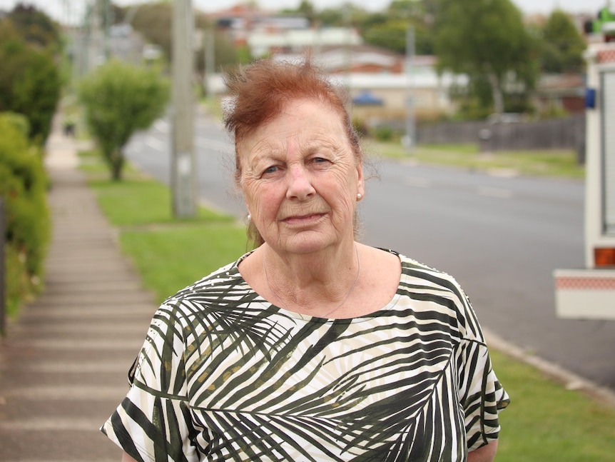 An older woman with red hair wearing a black and white shirt stands on a footpath in a suburban street.