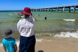A woman stands on the beach wearing a red hat and t-shit with 'SAVE OUR JETTY' handwritten on it. 