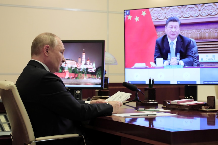 Man seated at desk speaks to another man on large television screen.