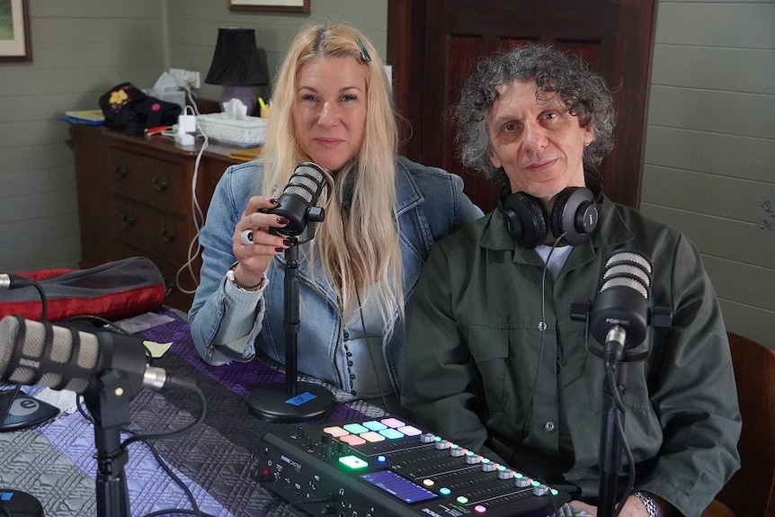 A blonde woman and a dark haired man sit behind a microphone with headphones around their neck