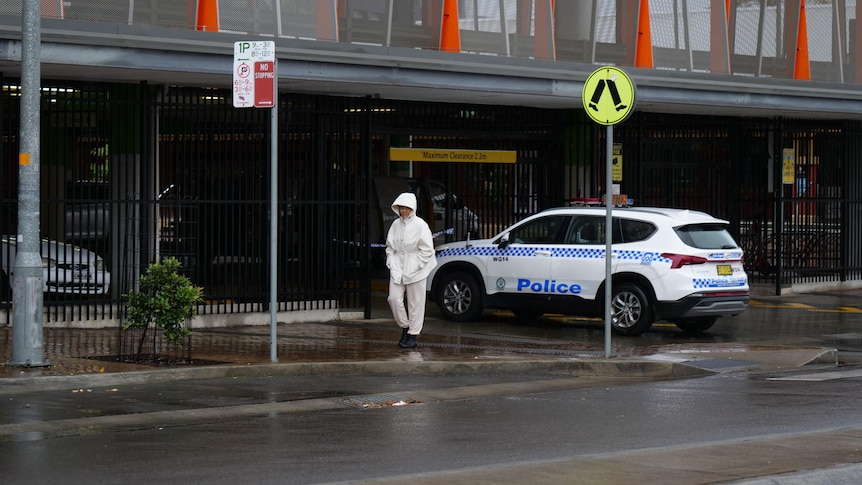 A person in a forensic suit walks near a car park on a rainy day.