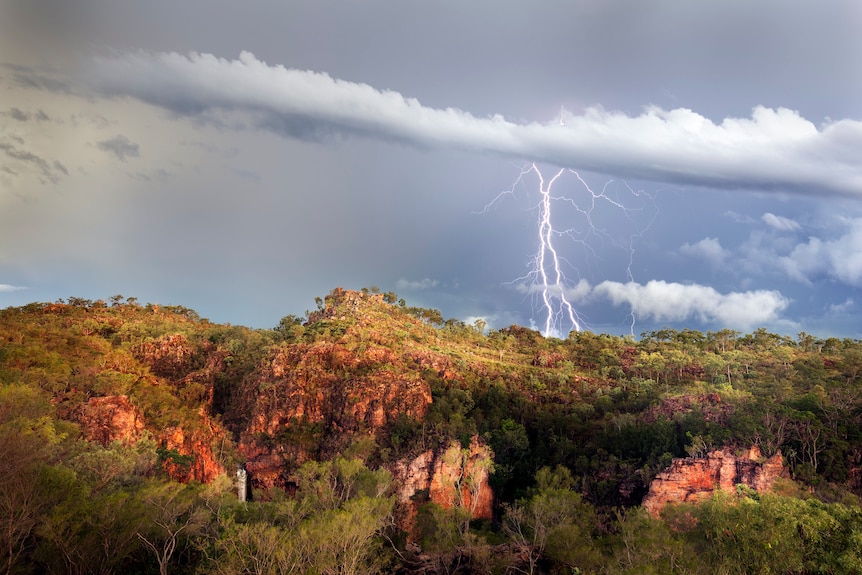 Fork lightening cracks through a grey sky over a red rocky, treed cliff face. 