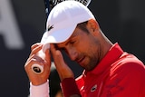 Novak Djokovic, wearing a white cap and orange shirt, wipes his brow during an Italian Open tennis match.