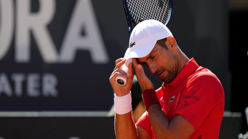 Novak Djokovic, wearing a white cap and orange shirt, wipes his brow during an Italian Open tennis match.