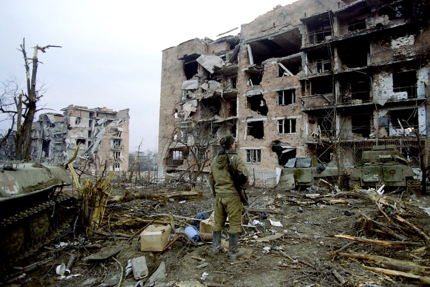 A soldier stands among the rubble outside a partially destroyed building. Entire walls collapsed, bricks and metal everywhere