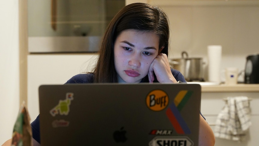 A woman with frustrated expression seen looking into laptop screen with blurred kitchen behind her.