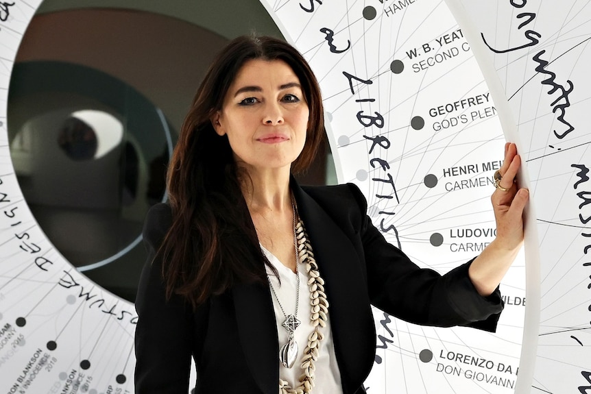 A woman with long dark hair, wearing a black blazer and white dress, standing in a spiral of white card printed with black text