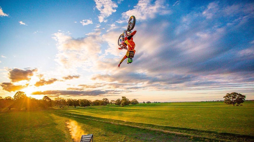 A motocross rider doing a mid-air back flip with the blue sky and clouds up above and green pasture below.