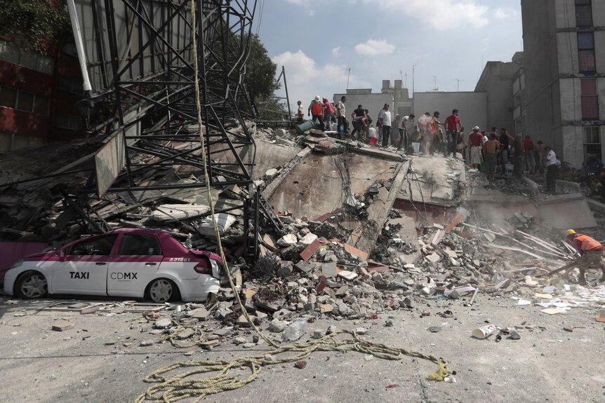A crowd of people stand on the dusty rubble of a building that has crushed a taxi.