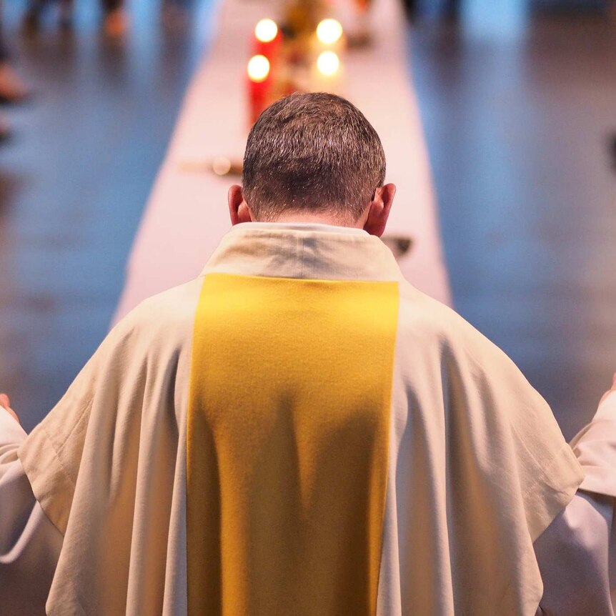 Photo of priest saying mass in church, back to the camera, facing towards congregation, arms outstretched, head bowed in prayer