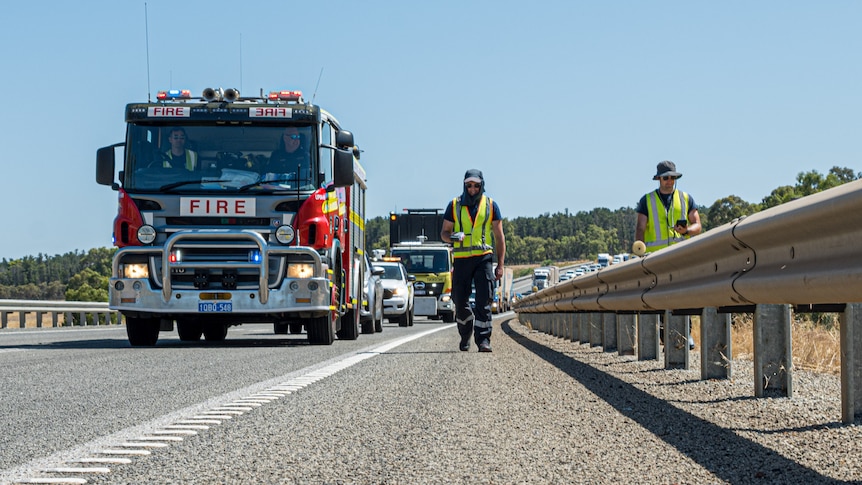 A fire truck driving and two people in high vis clothing walk along a busy highway.