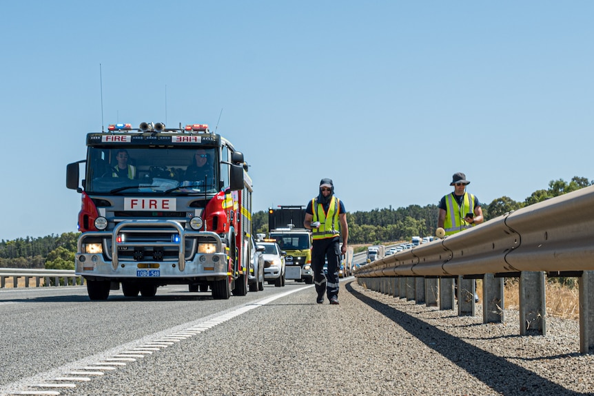 A fire truck driving and two people in high vis clothing walk along a busy highway.