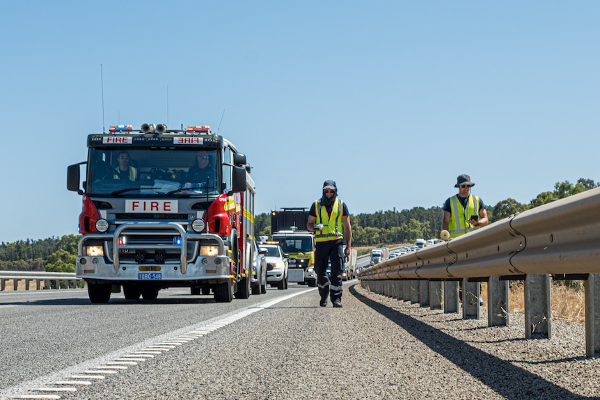 Un camion de pompiers conduit et deux personnes portant des vêtements haute visibilité marchent le long d'une autoroute très fréquentée.