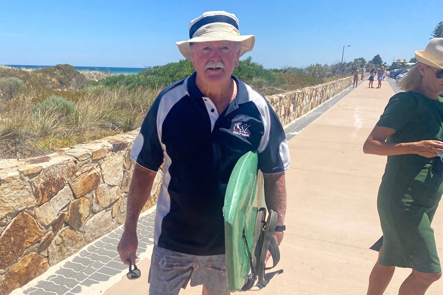 A man wearing a straw hat, navy shirt, shorts and carrying a green bodyboard stands in front of a beach